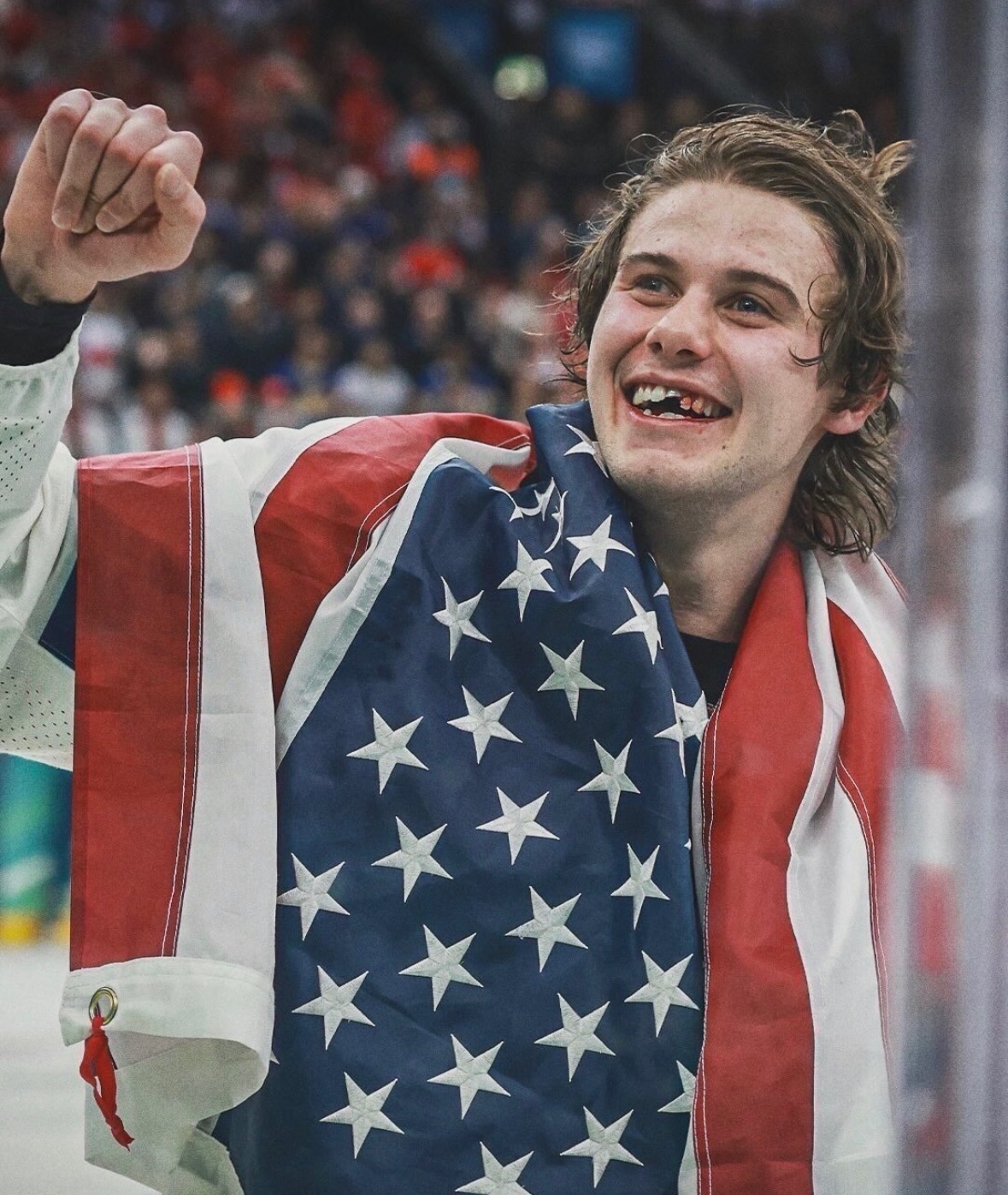 A man, Jack Hughes, smiles while raising his fist. He is wearing an American flag draped over his shoulders.
