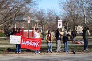 Protesters stand with signs along a street. One sign reads "Migration is not a crime" and another reads "Land back."