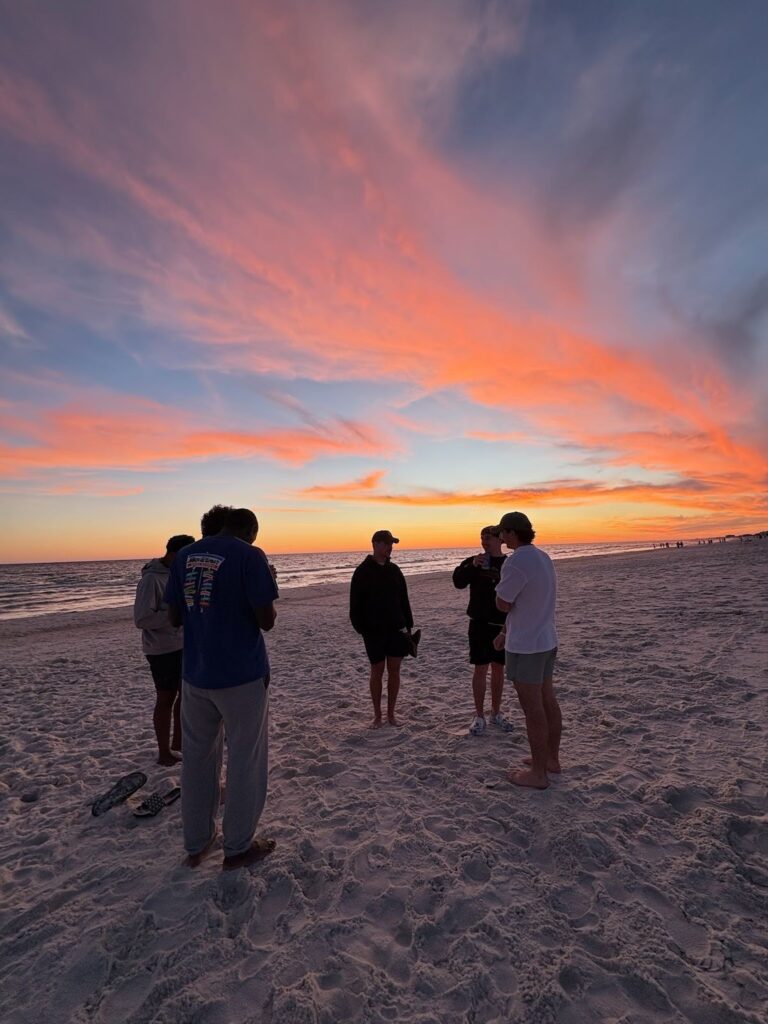 A group of people stands on the sand at a beach with a sunset in the background