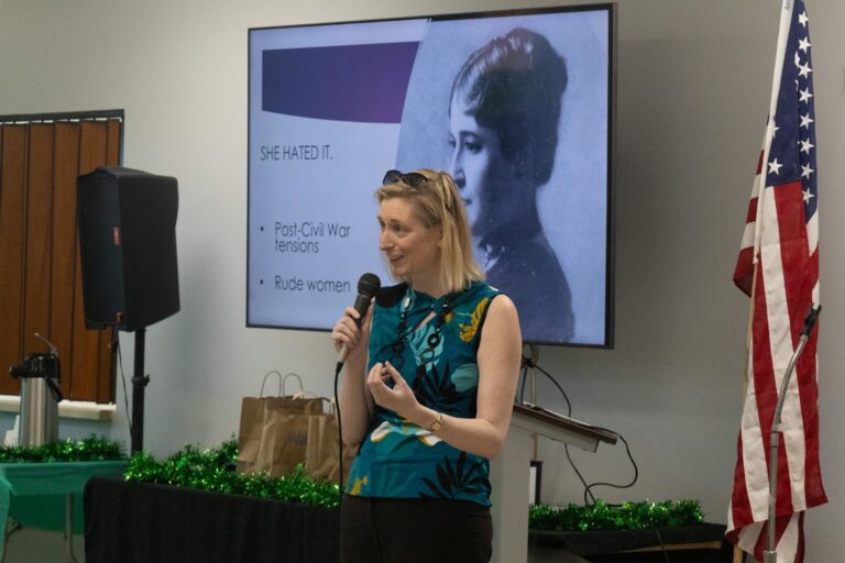 A person holds a microphone and is speaking. A slideshow is behind them.