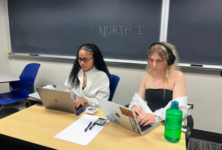 Two people sit side by side at a table looking at their laptops.