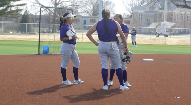 Three people stand facing each other inside a softball diamond