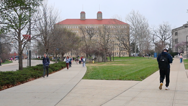 A person walks on a sidewalk with Fraser Hall at the University of Kansas in the distance