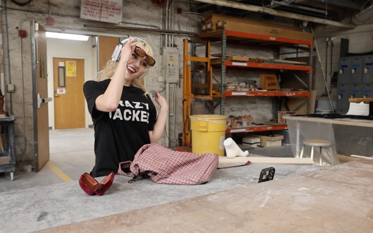 A girl wearing a black shirt, headphones, and sunglasses and films a video with a phone propped on a table. She is inside a room with shelves in the background.