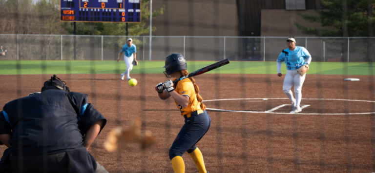 A softball player in yellow prepares to hit a softball pitched by a softball player in the right background wearing blue. An umpire in black is positioned to the left.