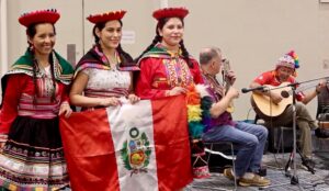 3 women in cultural attire hold a Peruvian flag. Performers play guitars in the background on the right.